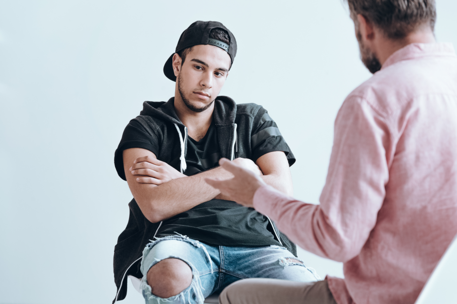 Teenage boy with hat sitting with arms crossed while adult talks to him
