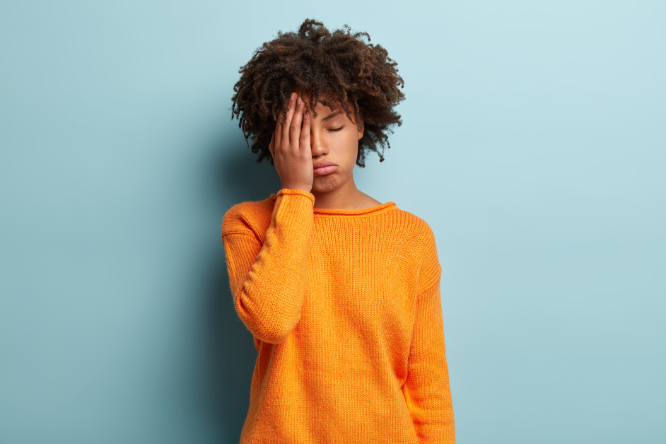 African American woman in bright orange sweater with hand on face looking tired against blue background
