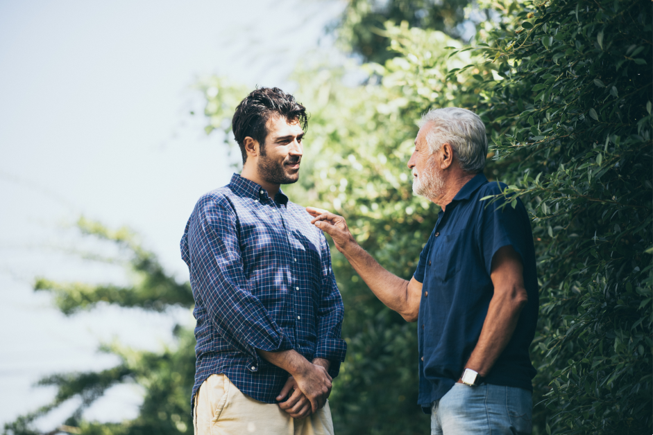 Man in blue shirt talk to older man in front of blue sky and trees